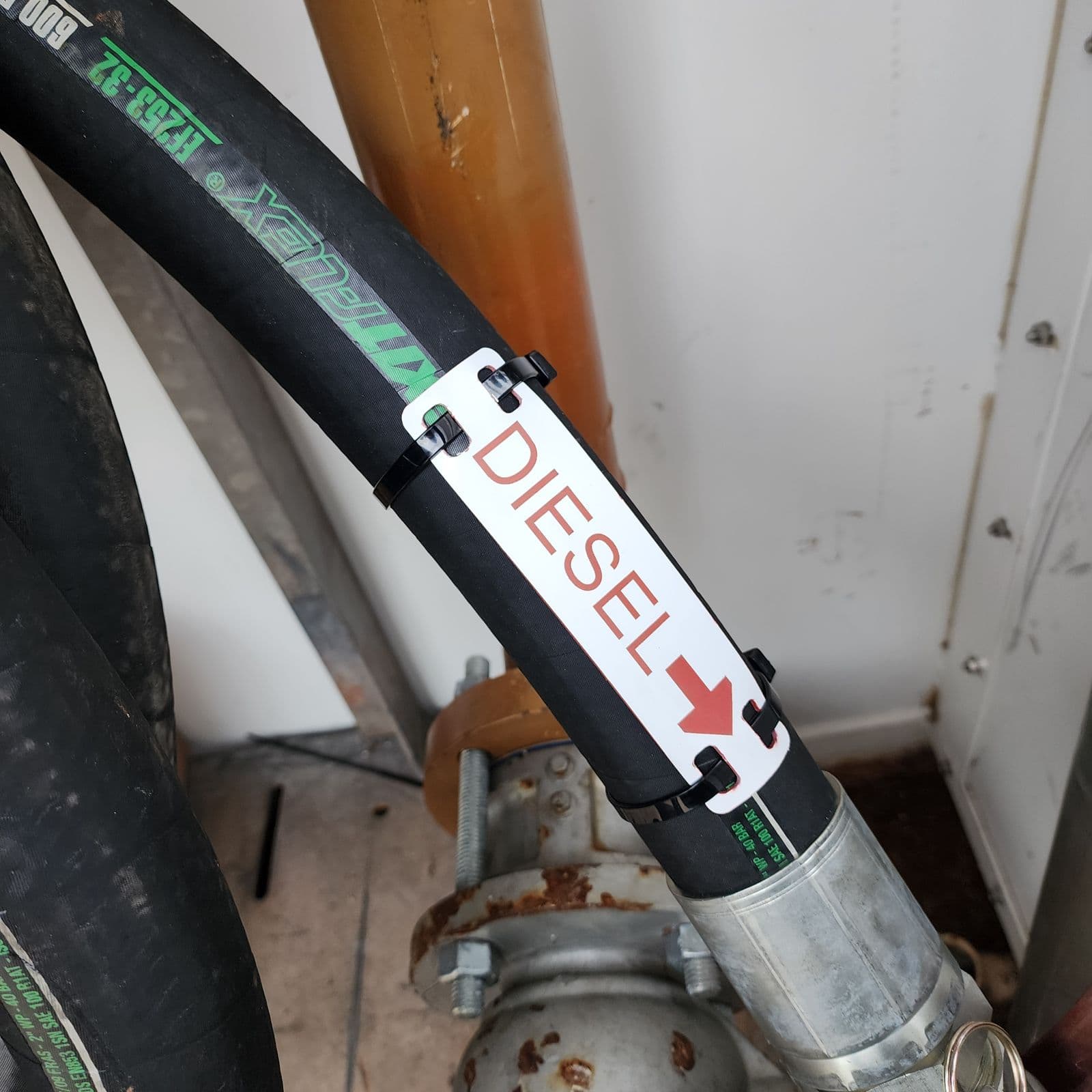 White traffolyte DIESEL hose marker with a red flow-direction arrow, cable-tied through slotted mounting holes to a black diesel supply hose outdoors