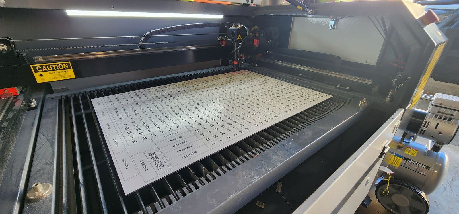 Laser engraving machine mid-cut on a dense white traffolyte sheet of numbered circuit labels