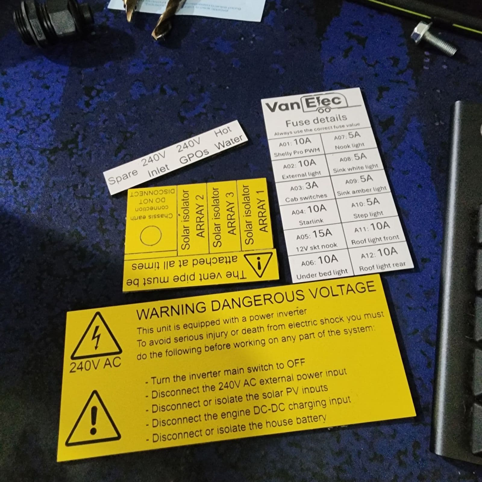 Set of van electrical fit-out labels on a workbench: a VanElec fuse detail card, solar isolator array labels, and a large yellow 240V AC Warning Dangerous Voltage sign